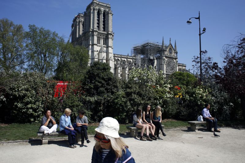 People sit in a park near Notre Dame cathedral, in Paris, Friday, April 19, 2019. Rebuilding Notre Dame, the 800-year-old Paris cathedral devastated by fire this week, will cost billions of dollars as architects, historians and artisans work to preserve the medieval landmark. (AP Photo/Thibault Camus)