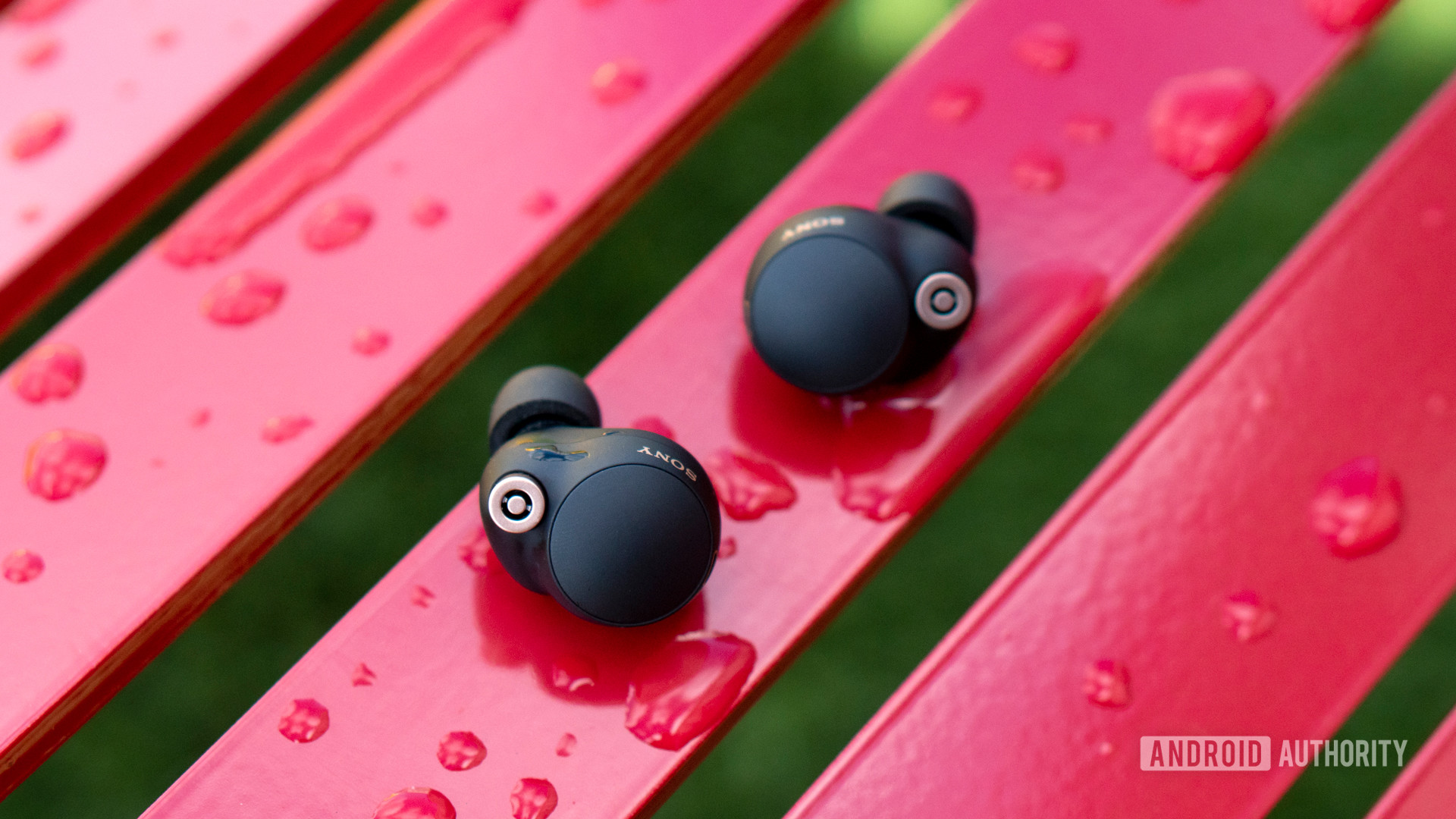 sony wf 1000xm4 review hero Sony WF-1000XM4 earbuds sitting outside of their case on a red bench covered with water droplets.