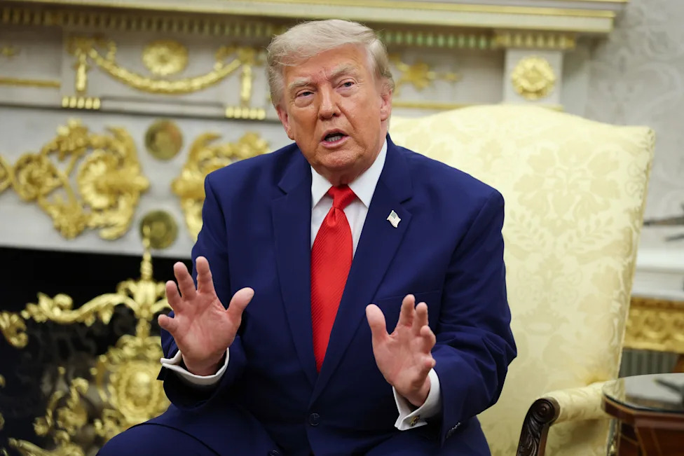 President Trump, in a blue suit, sits in a golden upholstered chair in the Oval Office of the White House. 