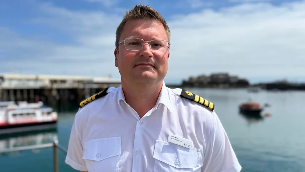 James Way is wearing his uniform, complete with epaulets, and is standing in front of the harbour in the sunshine. He has short dark blonde hair and is wearing apair of glasses.