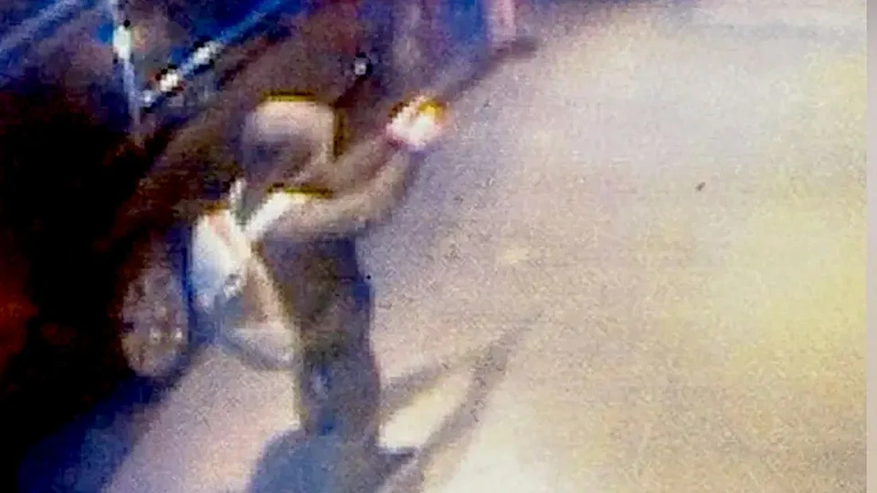 A hooded man wearing a backpack and aiming a gun with a silencer on a new york city sidewalk