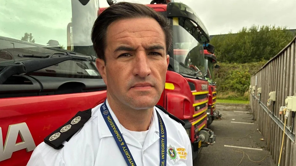 A man with dark hair and wearing a white shirt is standing in front of three red fire engines.