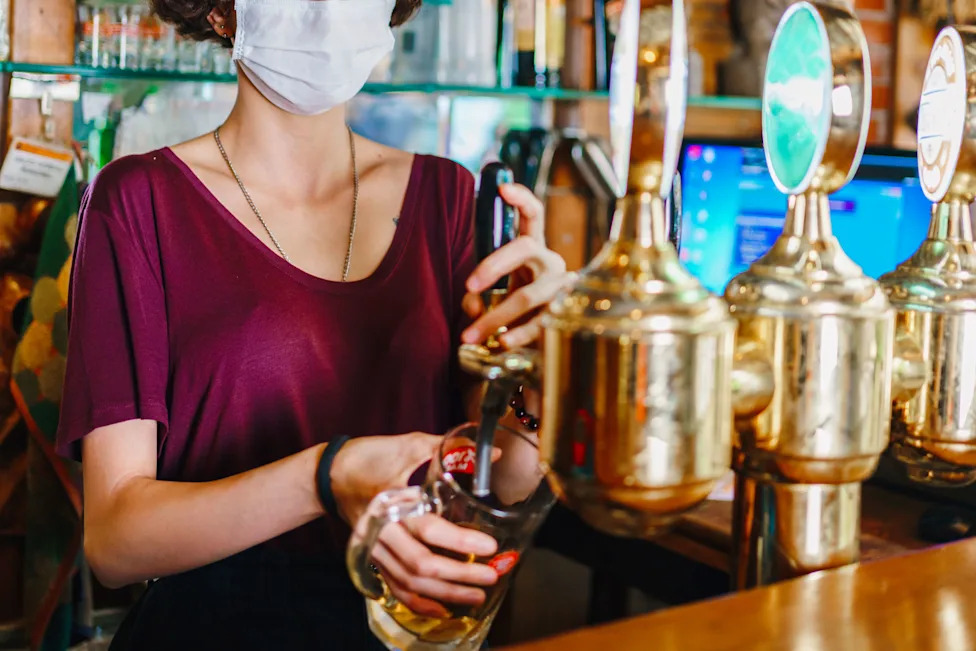 A stock photo shows a woman in a medical face mask pouring beer from a tap. 